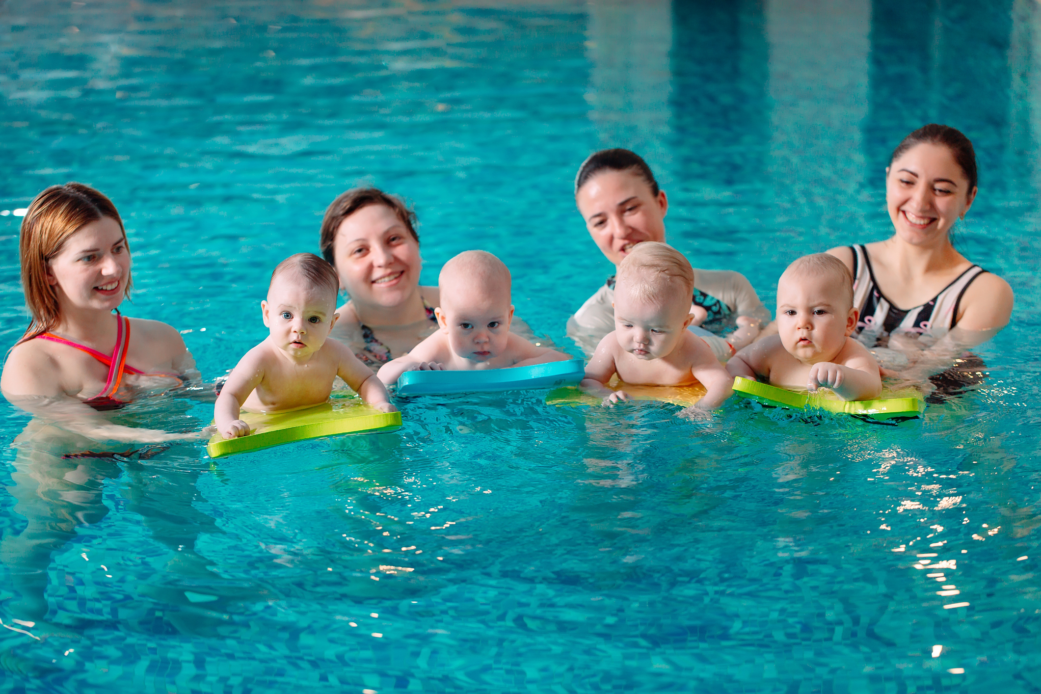 A group of mothers with their young children in a children's swimming class with a coach.